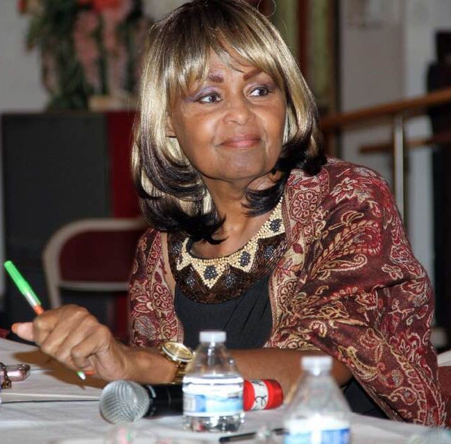 Woman with a brown wig, holding pen, at a table with microphone and water bottles.