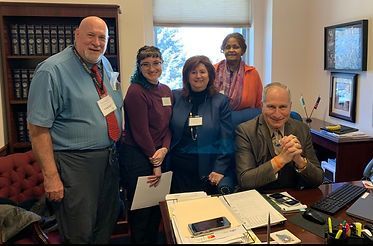 Five people in an office setting: three standing, two seated. They're near a desk with papers, a phone, and a computer.