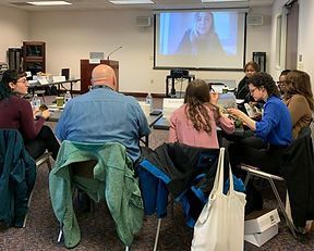 People seated around a table in a meeting room with a projected video call. Several are engaged in a discussion.