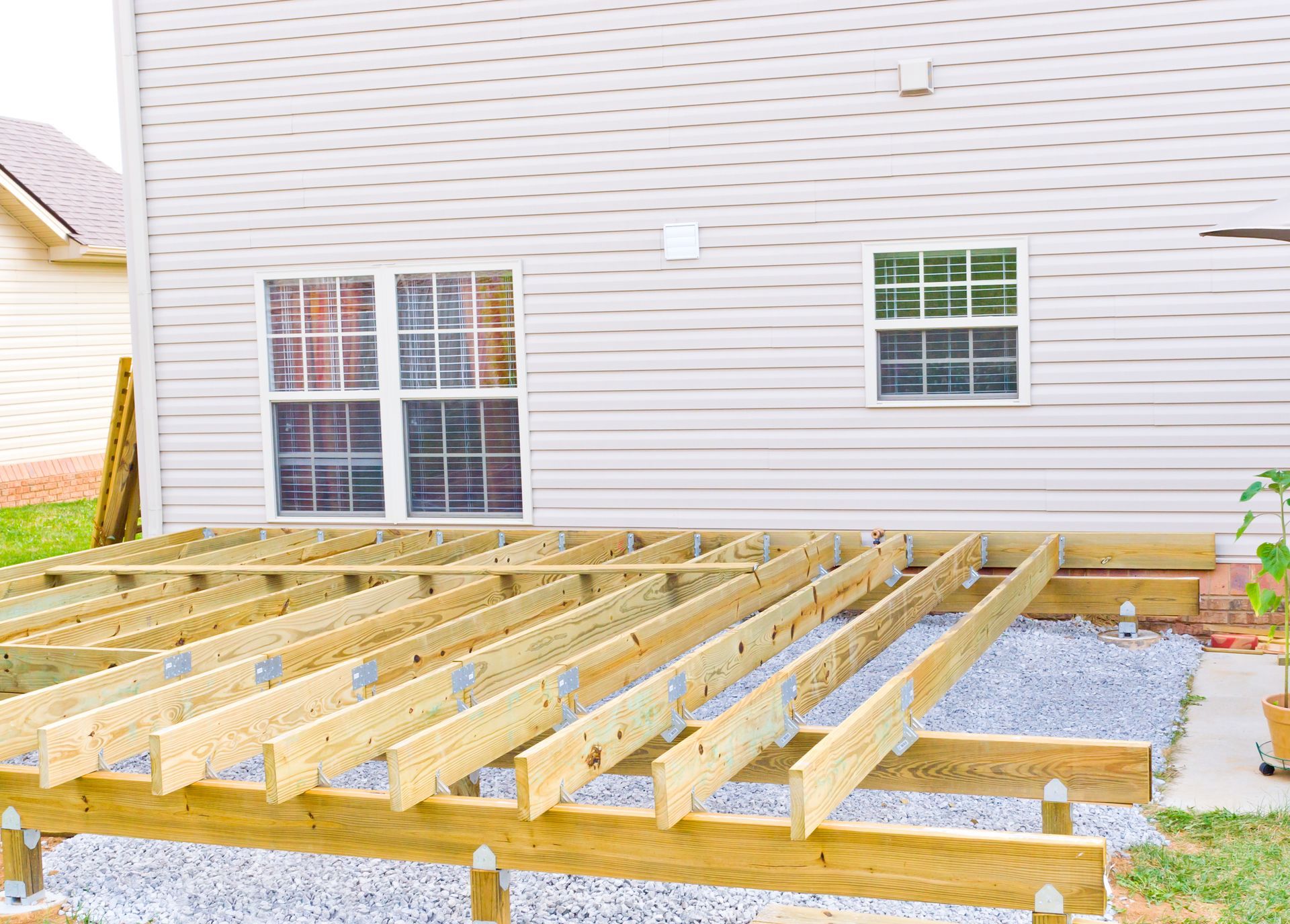 Wooden deck under construction next to a light-colored house with windows and a gravel foundation.