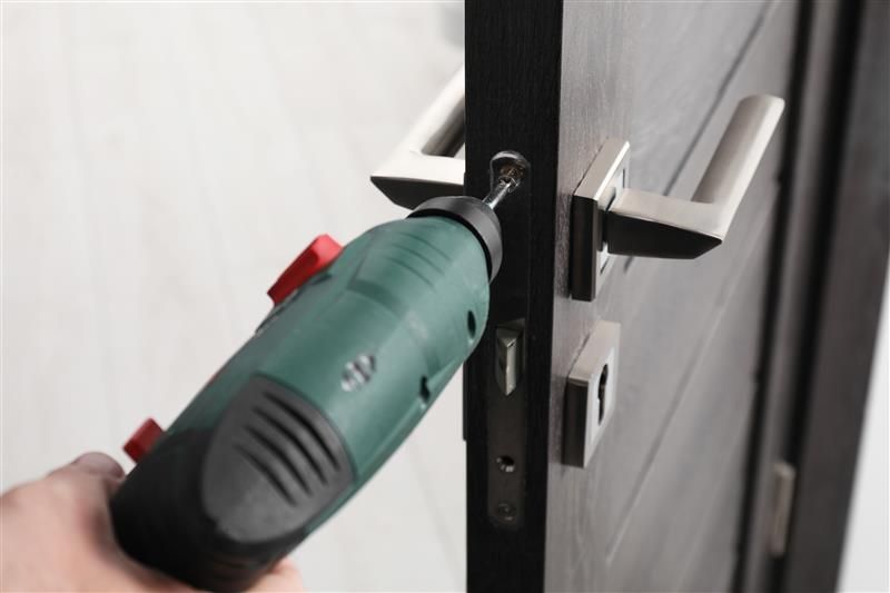 Person using a power drill to install a silver door handle on a dark wooden door.