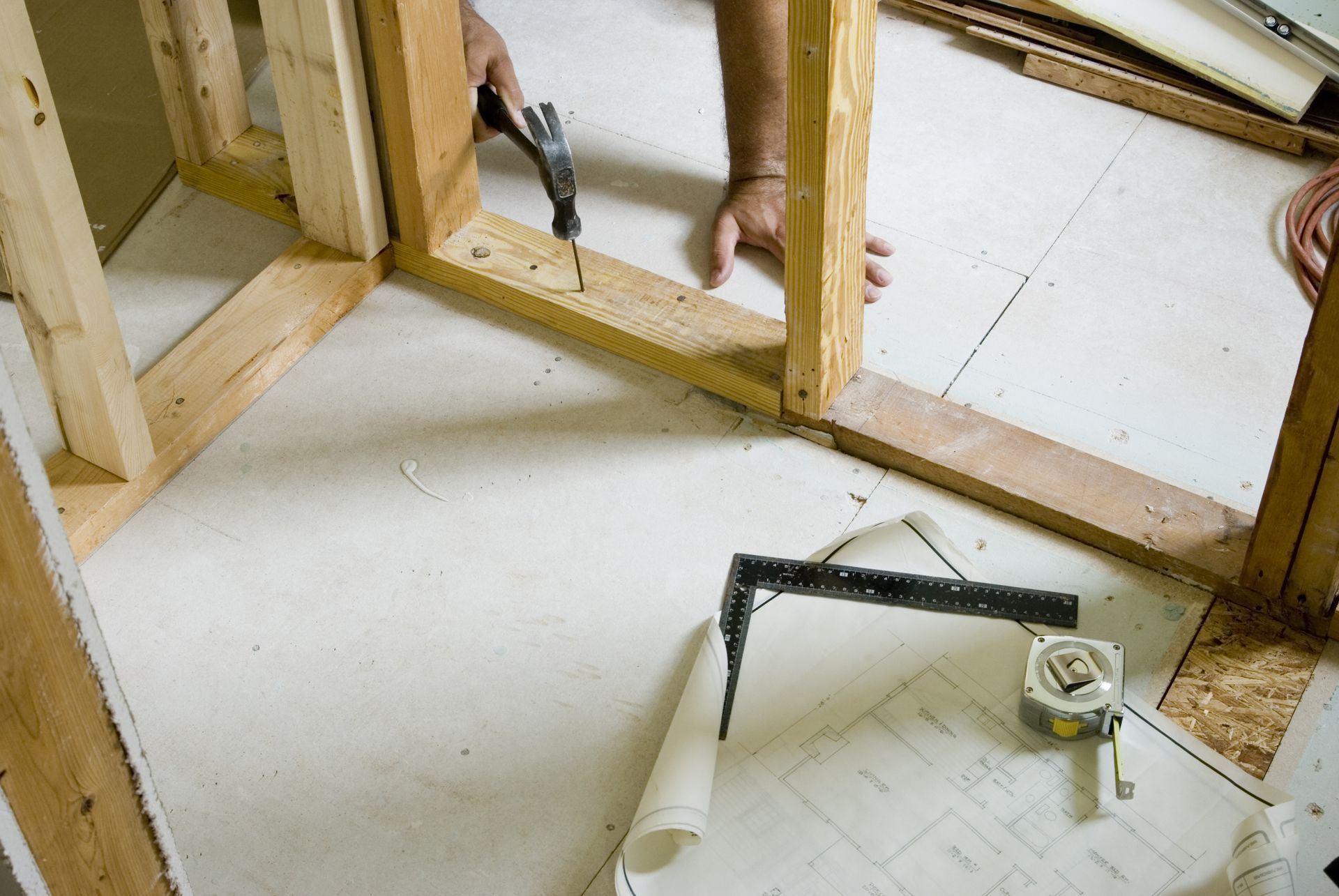Person constructing a wooden frame with a drill. Tools and blueprints are nearby on the floor.