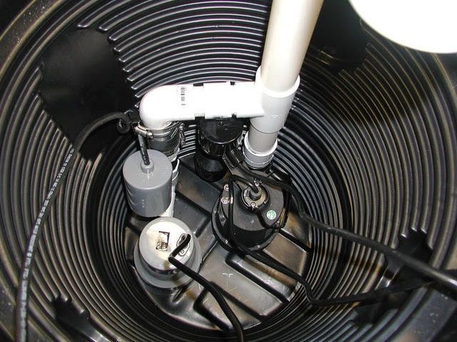 Black sump pump system inside a dark, corrugated plastic basin. Features pumps, floats, and PVC pipe.