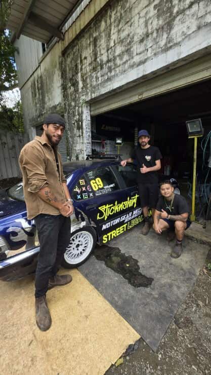 Three men pose with a blue race car in front of a garage. Two stand, one squats. The car has sponsor decals.
