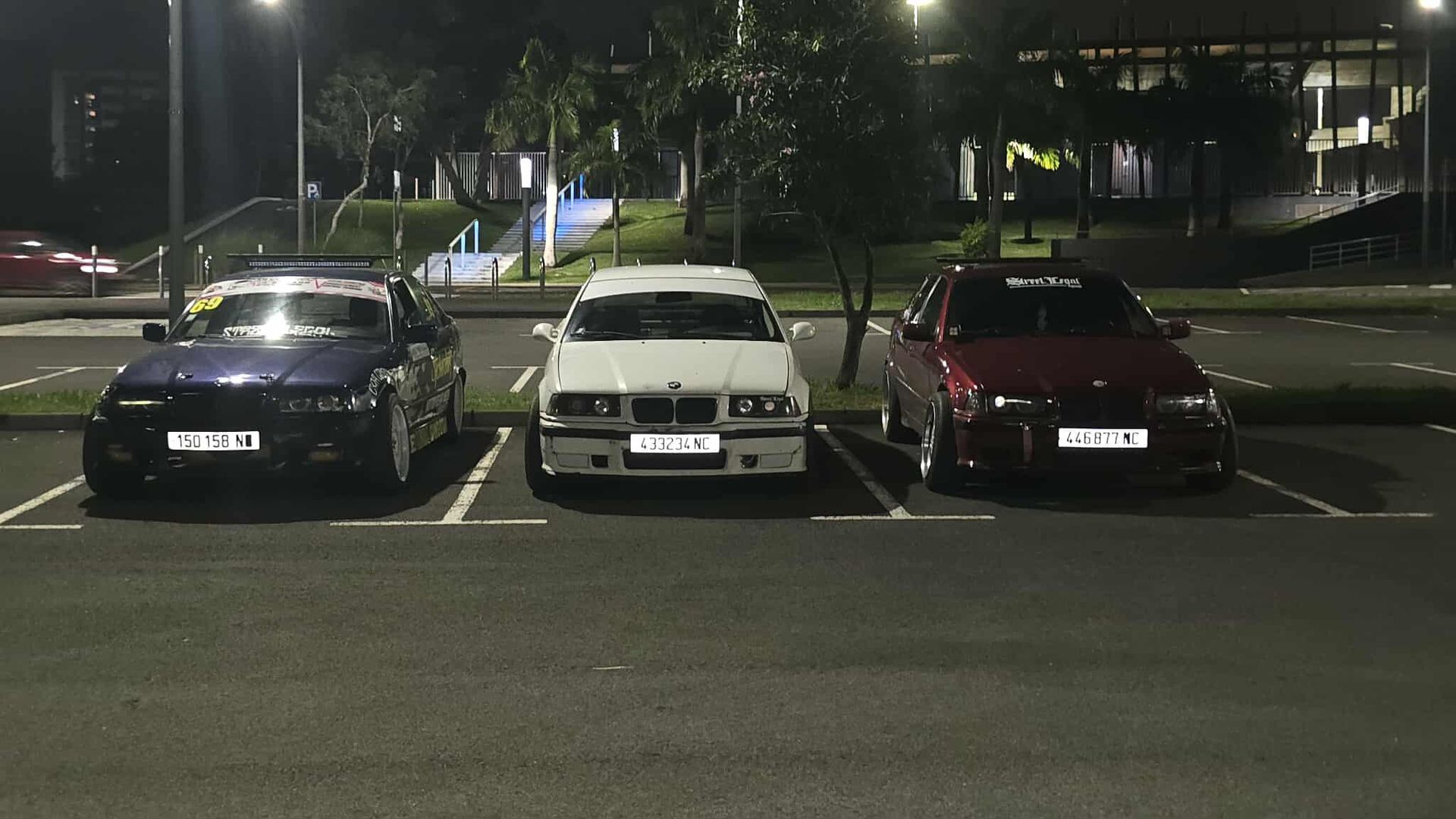 Three cars parked in a dimly lit parking lot at night: dark blue, white, and burgundy.