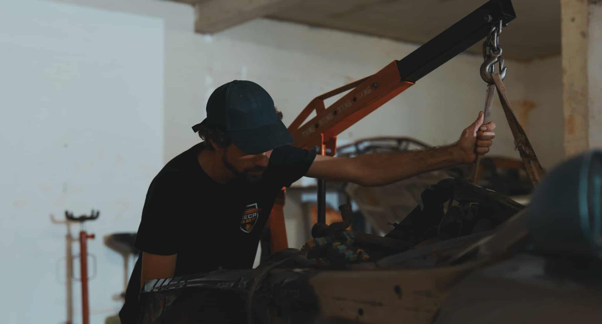 Man in black shirt and cap using a crane to lift a car engine in a garage.