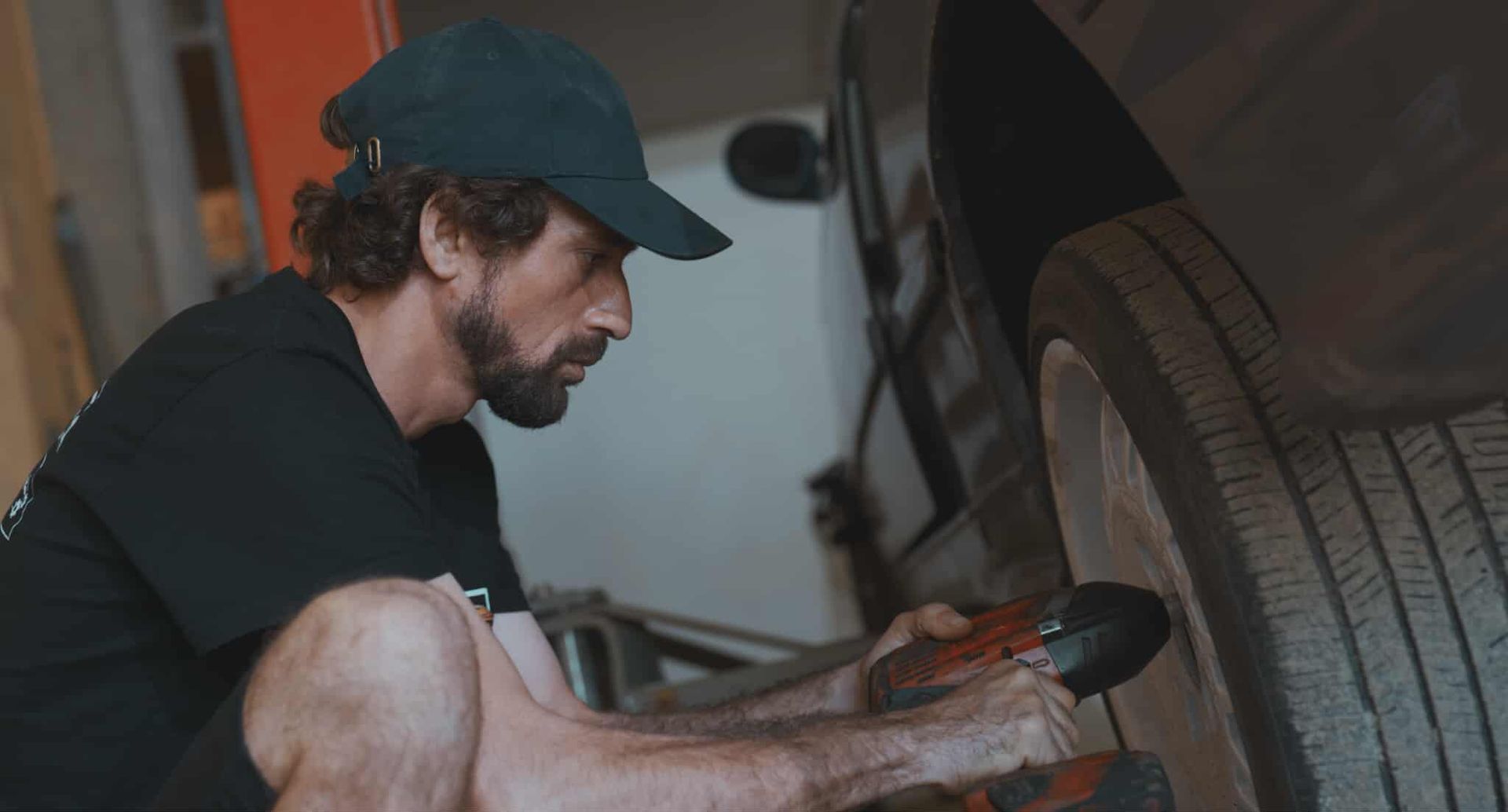 Man in a cap, working on a car tire in a garage, using a tool.