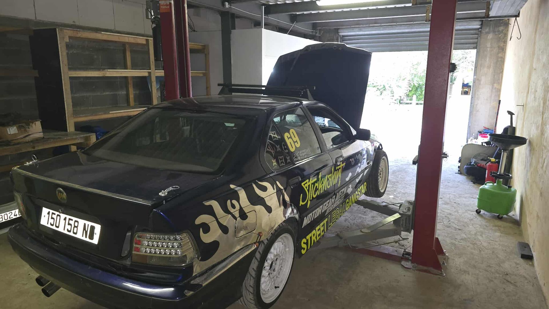 Dark blue car on a lift in a garage, hood open. White wheels, graffiti decals, and red lift posts.