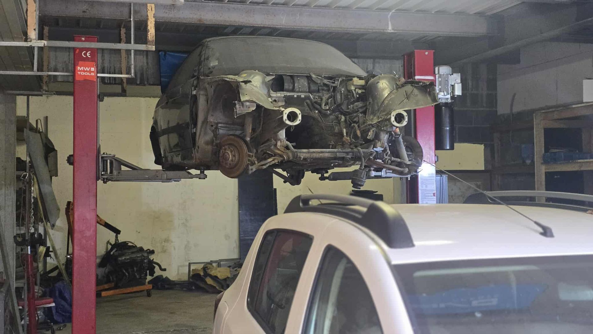 A disassembled car raised on a lift inside a garage. Another car is parked nearby.