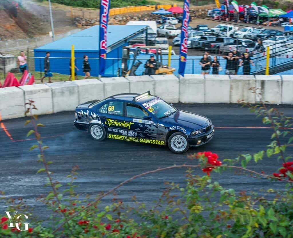 Blue car drifts on a track, smoke rising. People watch by a barrier with Red Bull banners.
