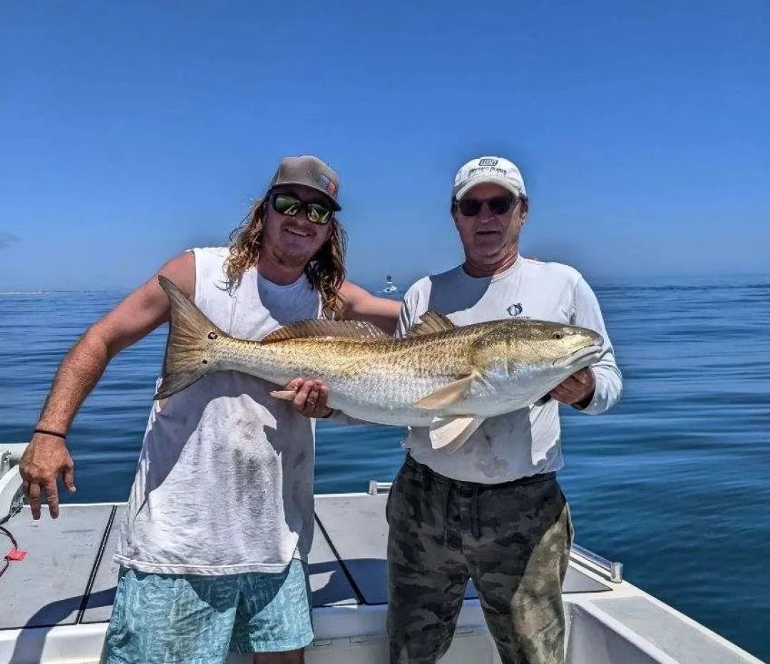 Two men on a boat holding a large redfish, smiling. Sunny day, blue water.