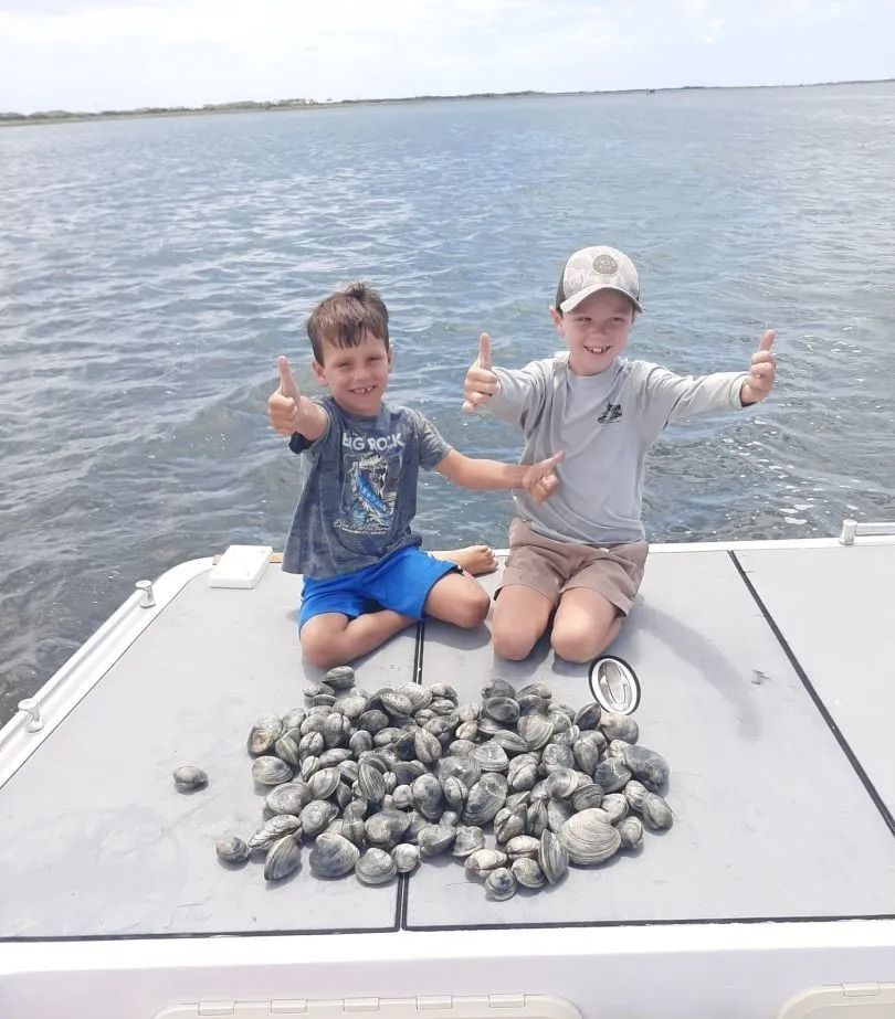 Two boys on a boat smile, giving thumbs-up, next to a large pile of clams. Blue water and sky in the background.
