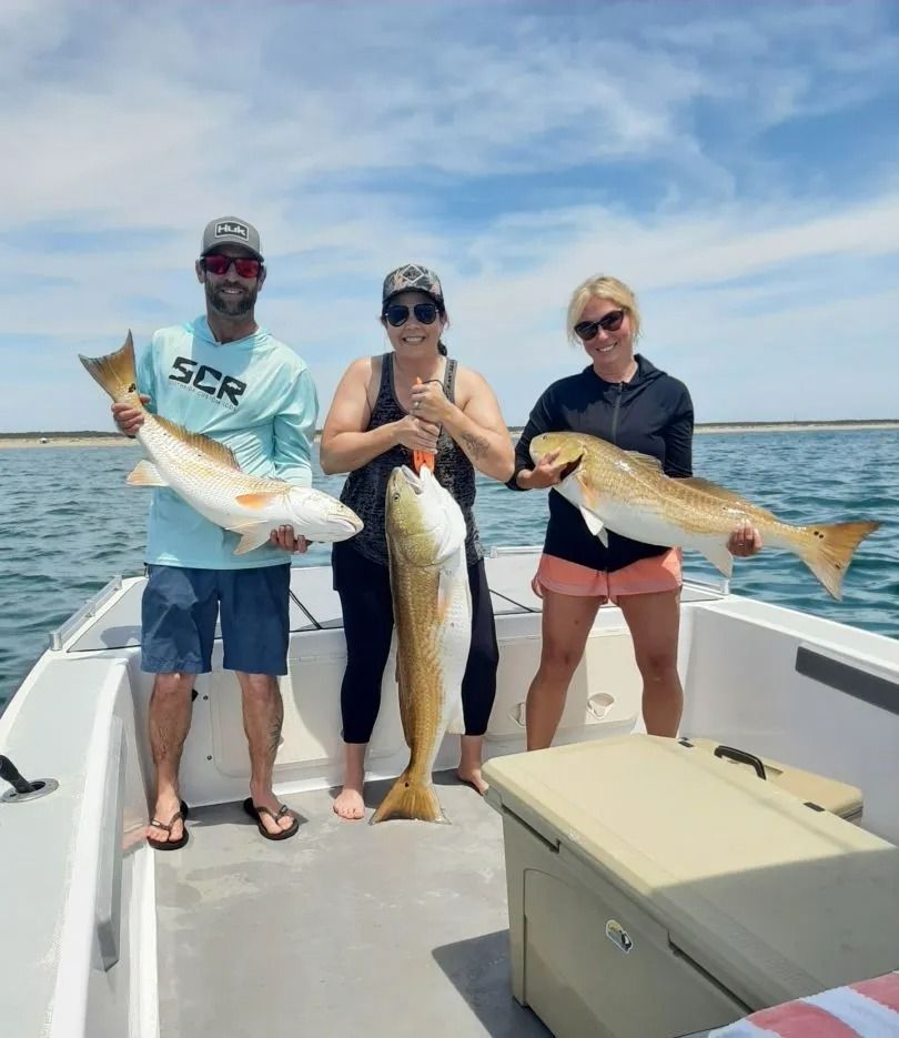 Three people on a boat hold up large redfish they caught in the ocean on a sunny day.