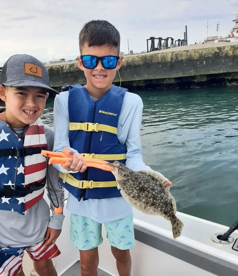 Two boys on a boat smile, holding a fish they caught, wearing life vests, by a dock.