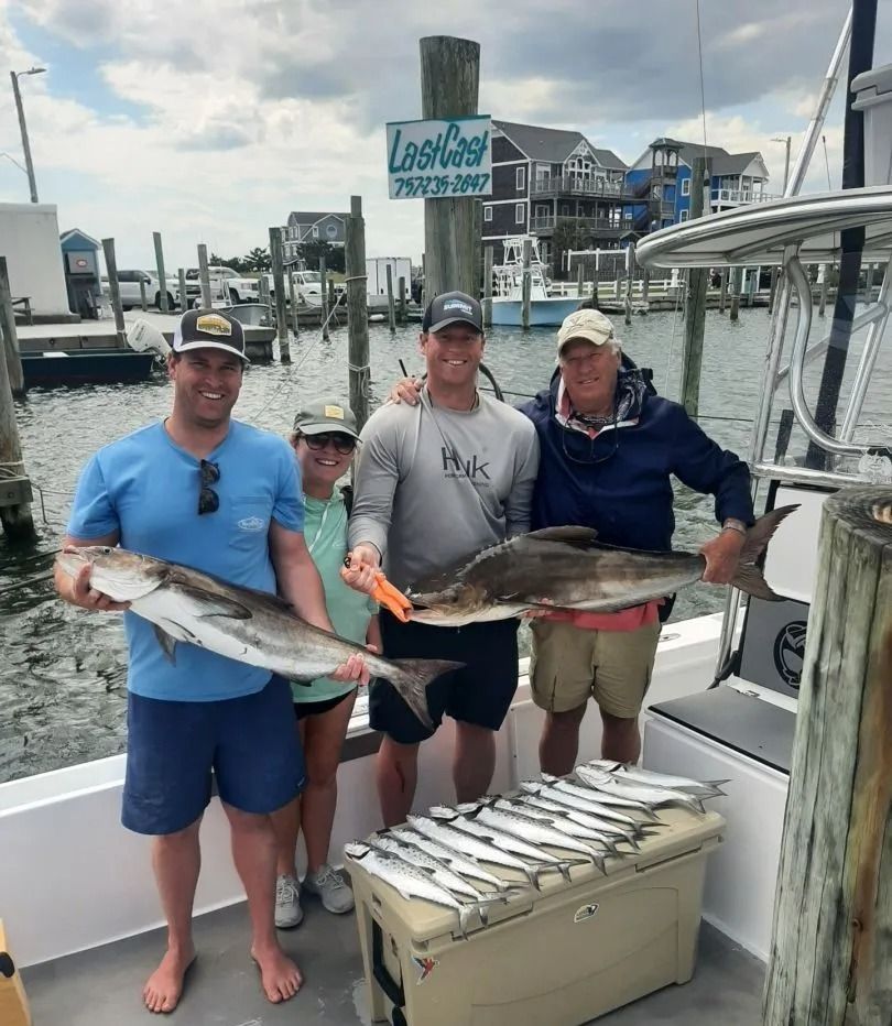 Four people on a boat proudly display caught fish. A cooler holds several smaller fish. Coastal setting, sunny.