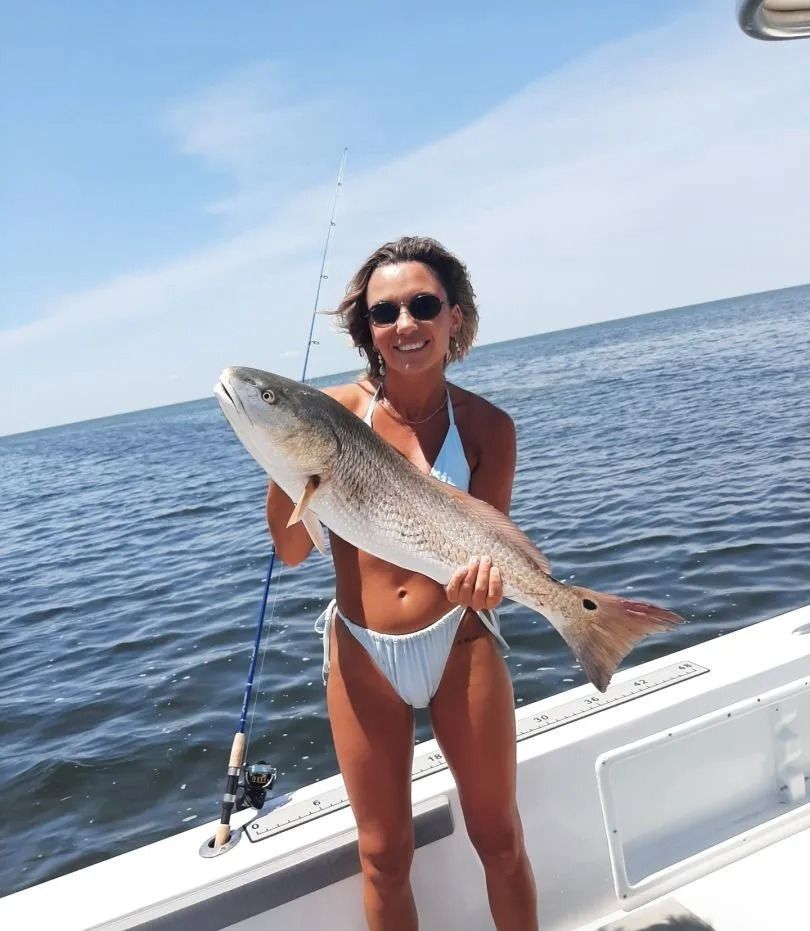 Woman in a bikini on a boat holds a large redfish, smiling, with fishing rod. Blue water and sky.
