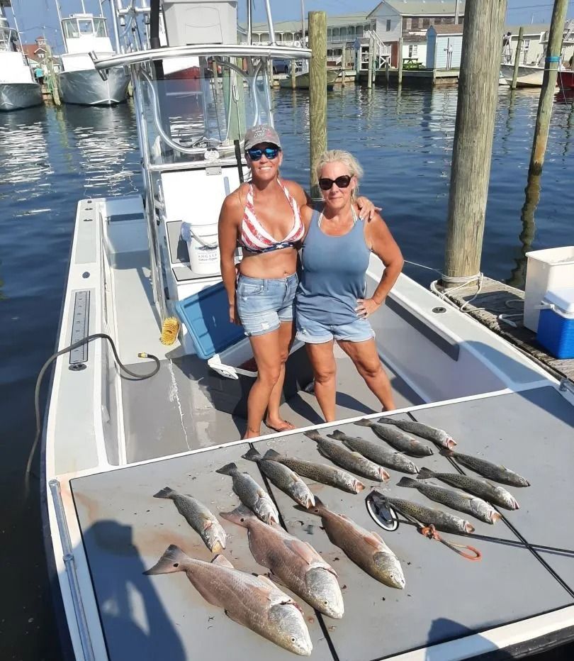Two women pose on a boat with caught fish. They are smiling, in summer clothes, at a dock.