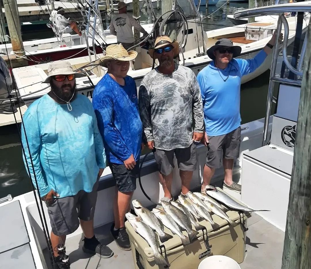 Four men on a boat, smiling, holding fishing rods, next to a cooler with caught fish.