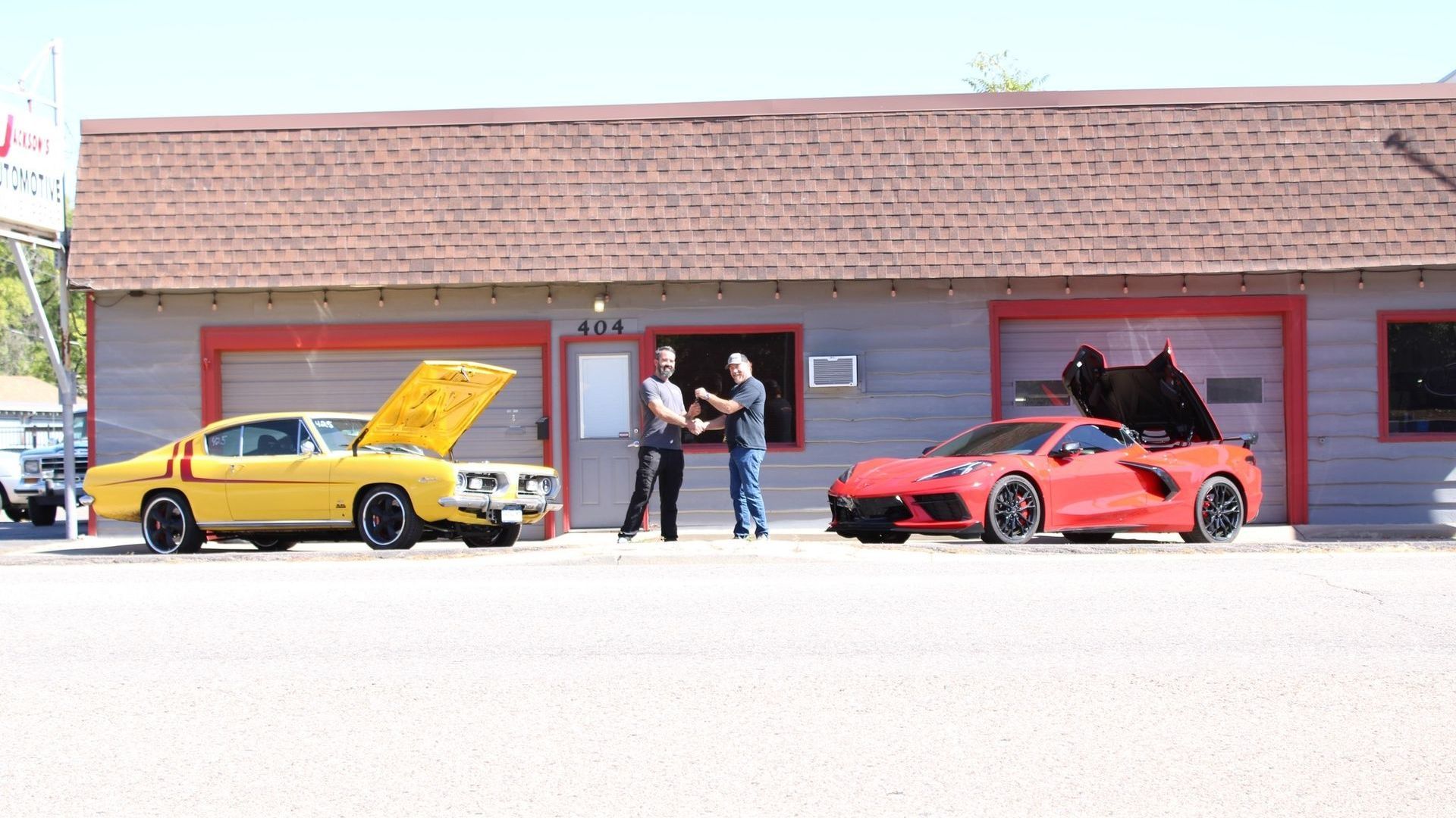 Two men are shaking hands in front of a building with two cars parked in front of it.