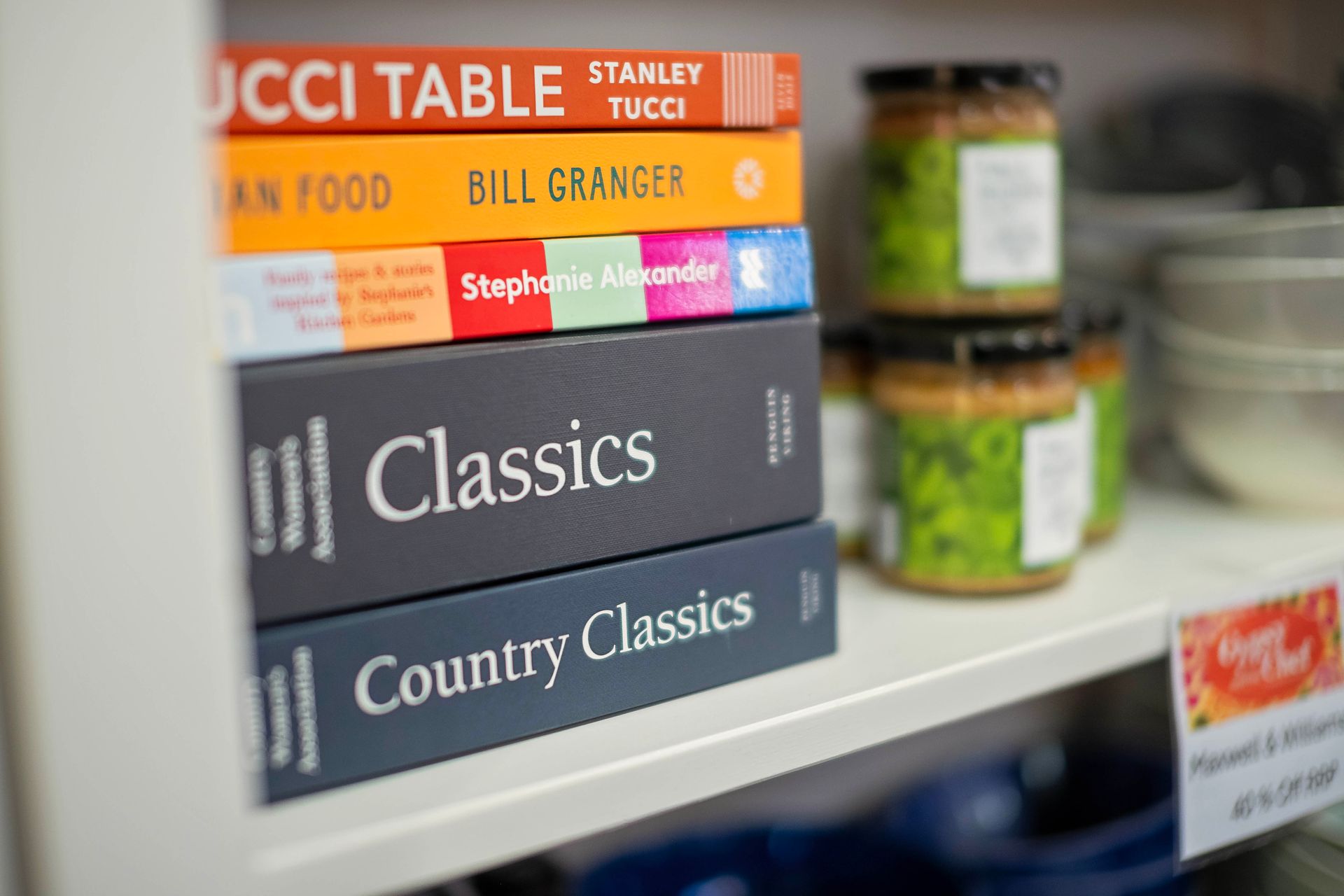 Cookbooks stacked on a shelf, with jars of food and bowls in the background.