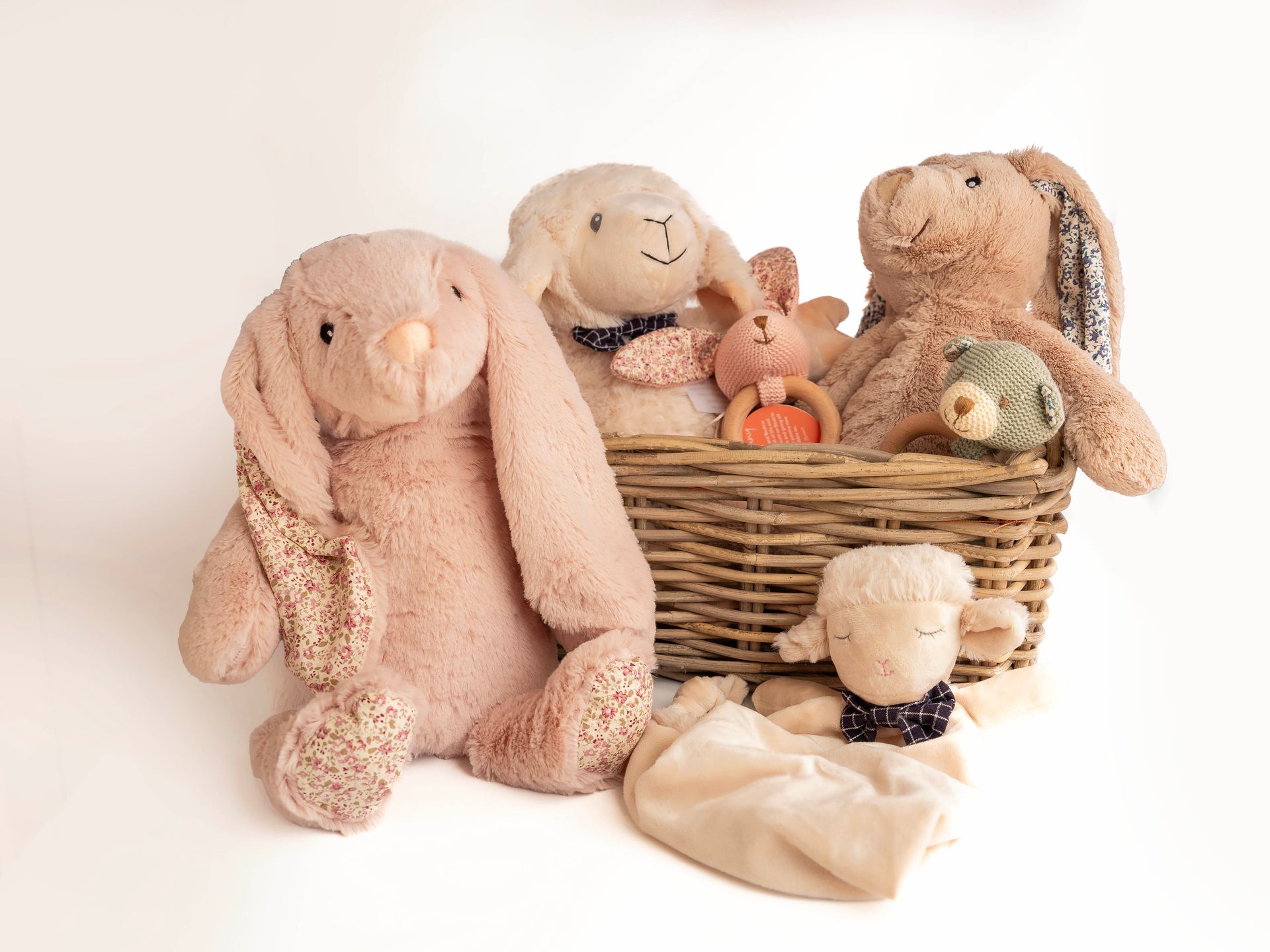 Collection of plush toys, primarily bunnies and lambs, in and around a wicker basket. Neutral colours, on white background.