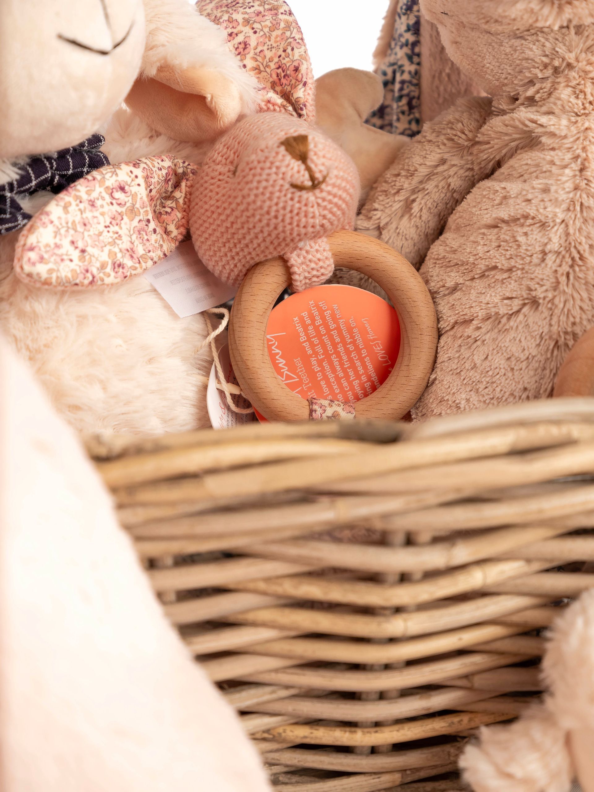 Wicker basket filled with soft plush toys in pastel colours, including a knitted bear, a wooden teether.