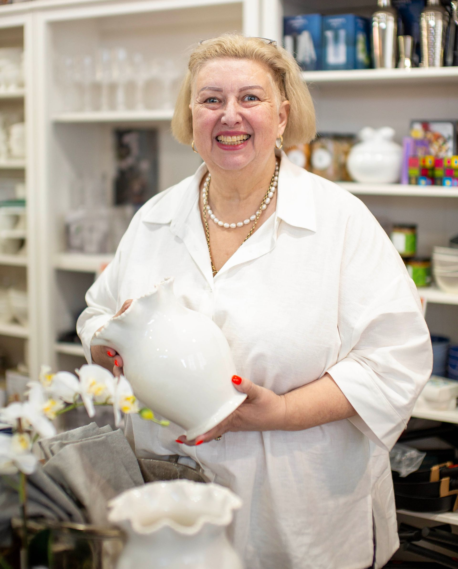 Woman in white shirt holding a white pitcher, smiling, in a shop with shelves of items.