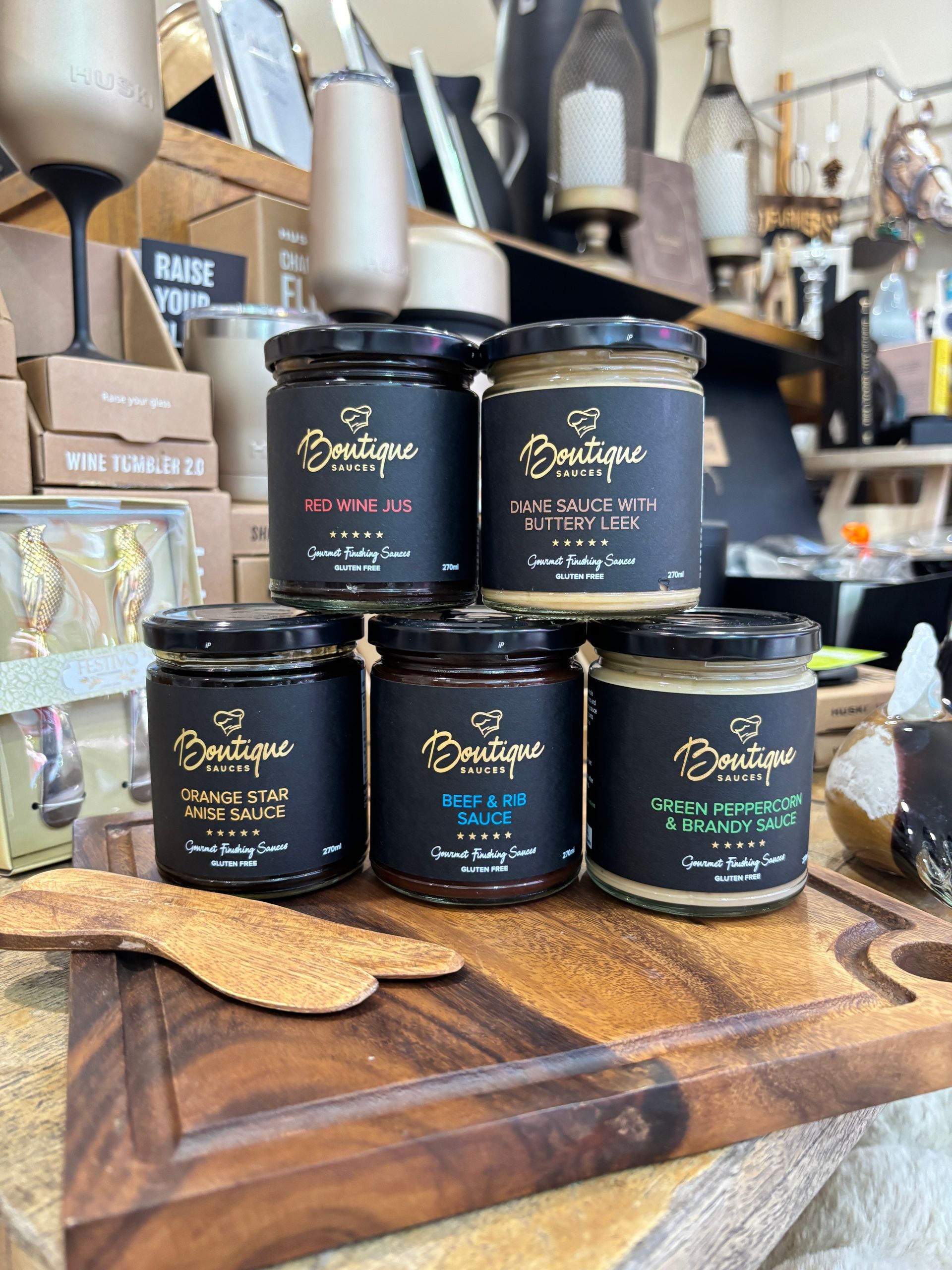 Five jars of gourmet food products on a wooden cutting board in a retail setting.