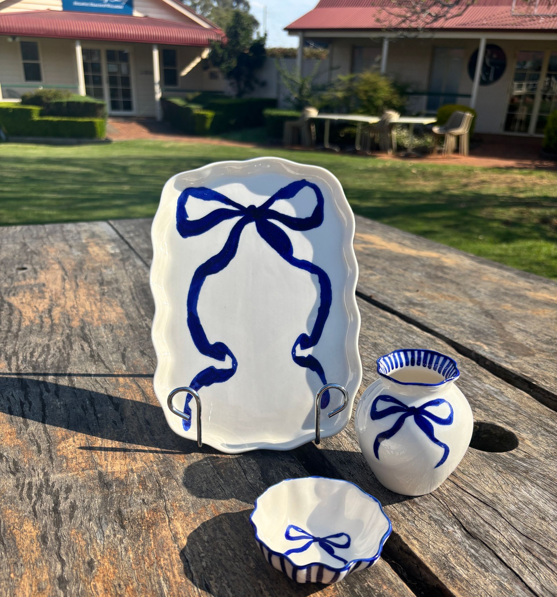 White ceramic dish, bowl, and vase with blue ribbon design, set on a wood table, backyard setting.