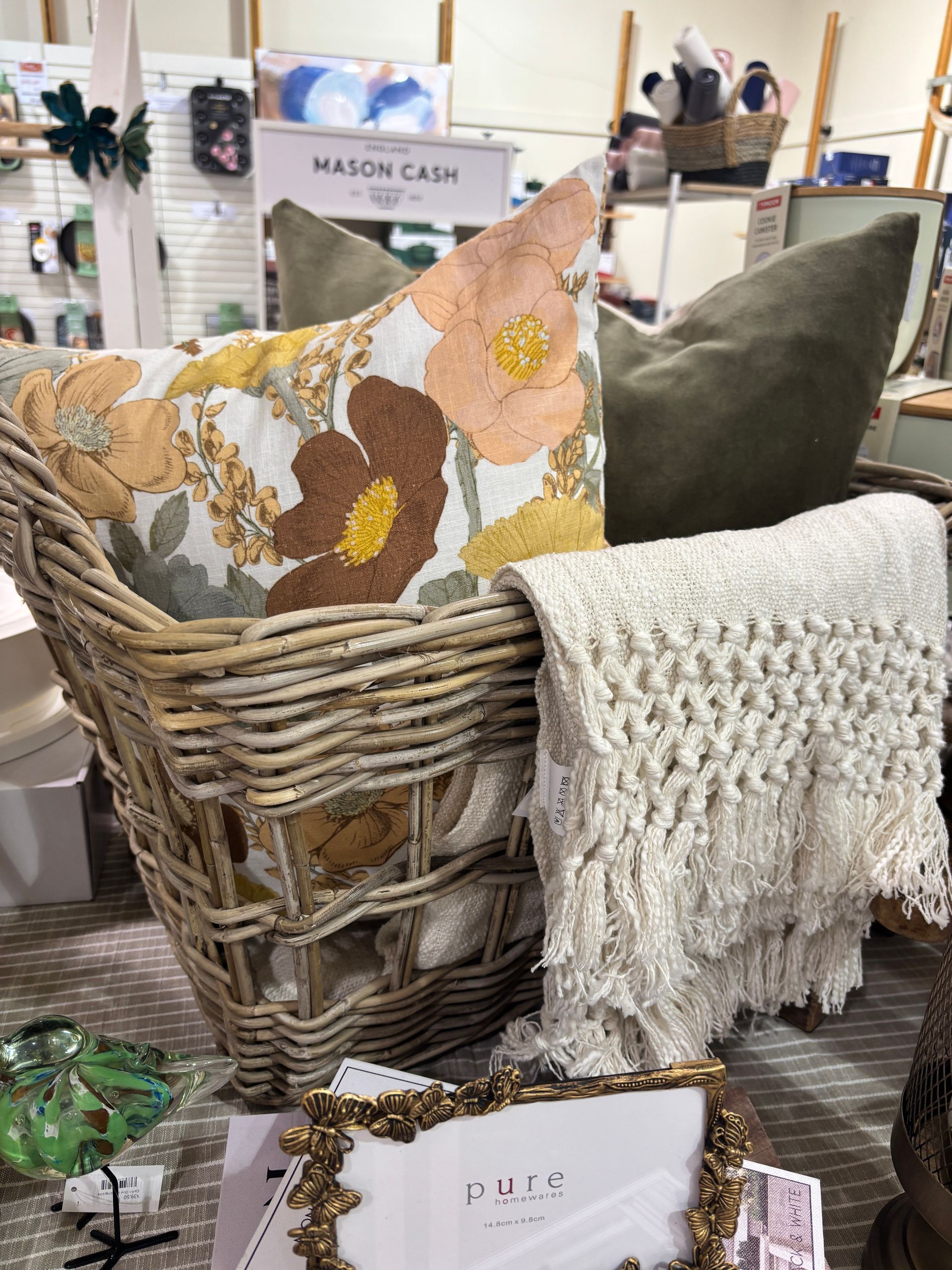 Wicker basket with floral pillow, green pillow, and fringed throw. Frame in front.