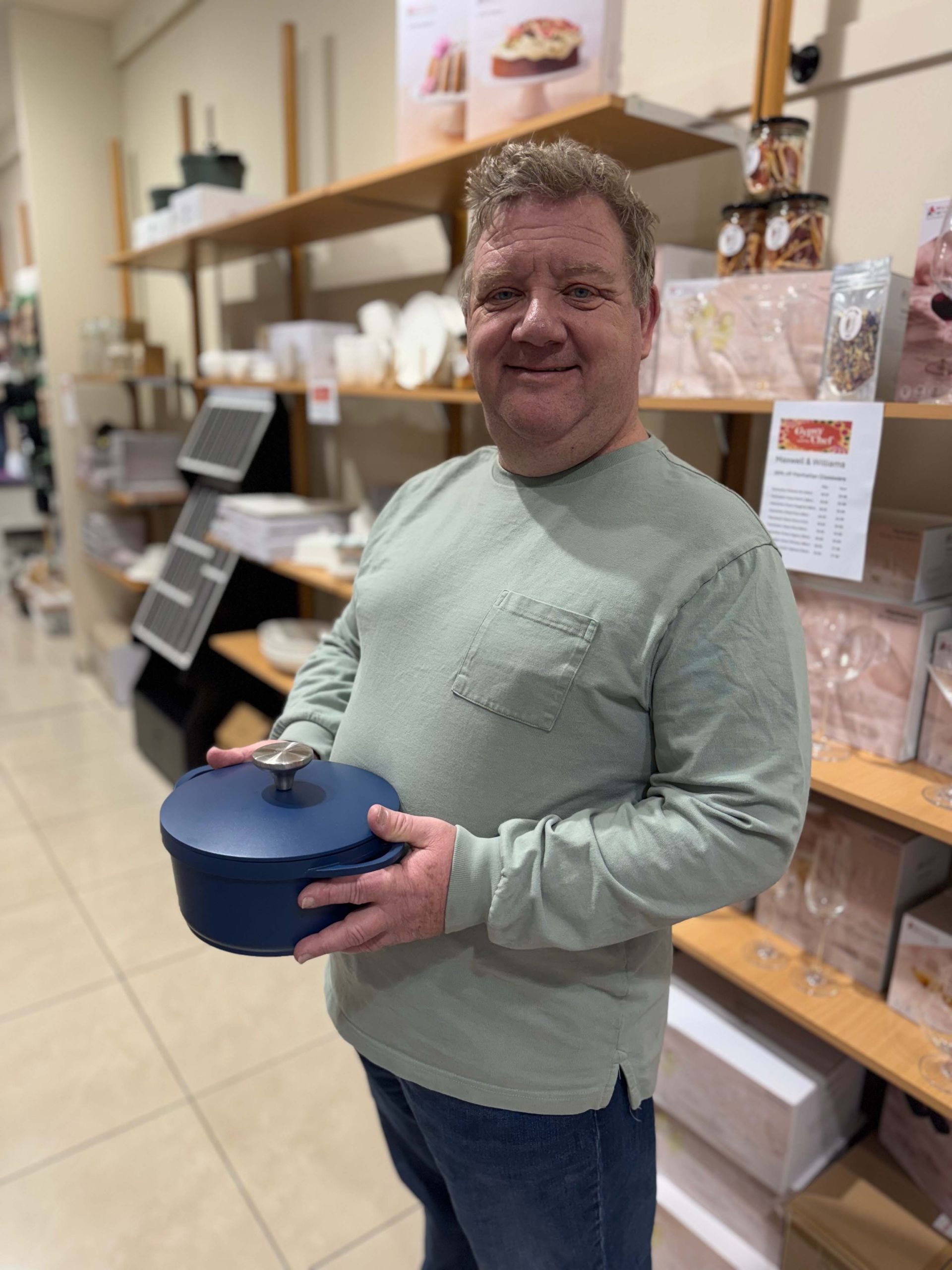 Man holding a blue pot in a store. He wears a green shirt, smiles. Shelves with items are in background.