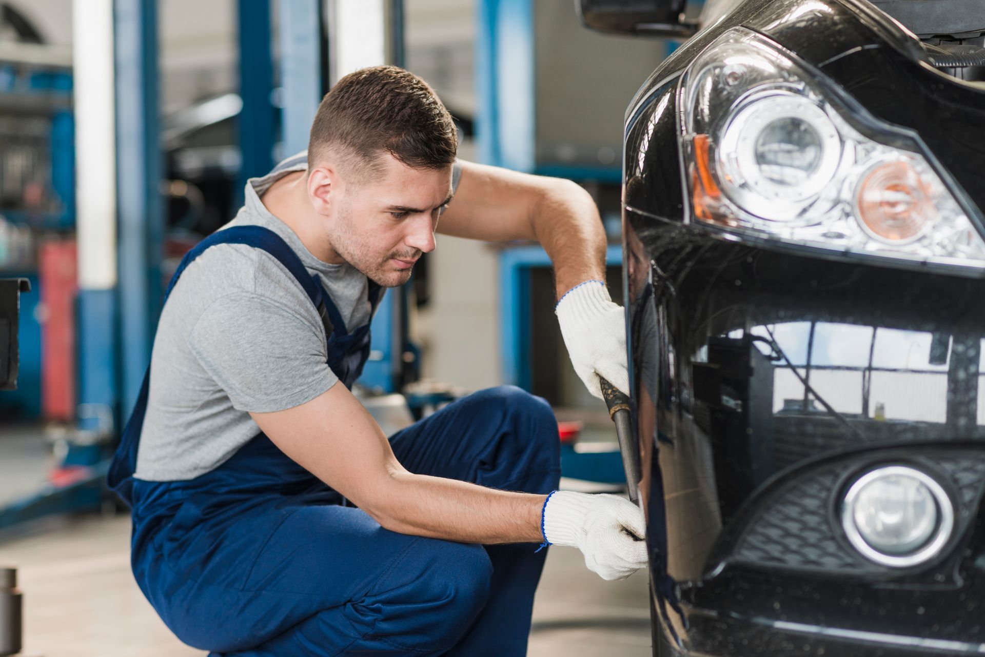 Mechanic in blue overalls works on a black car in a garage, wearing gloves.