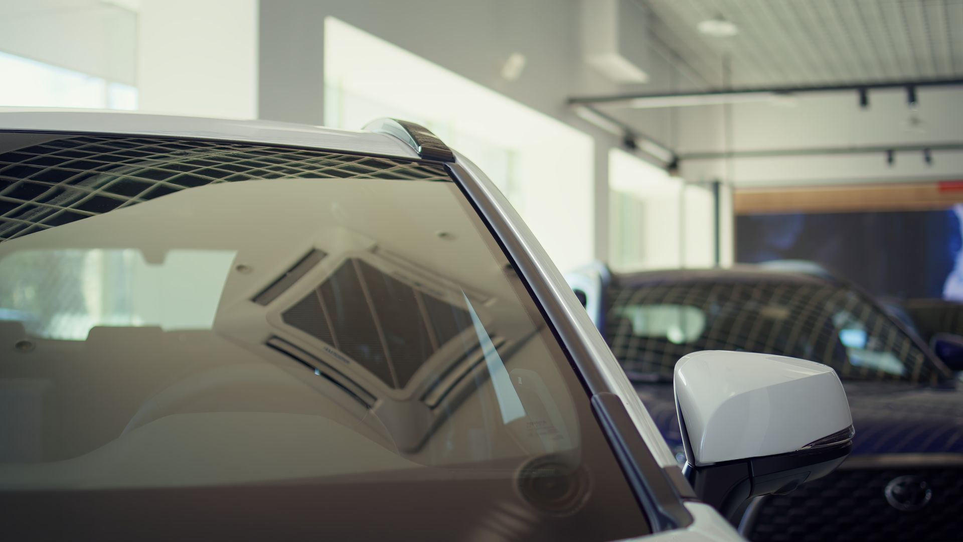 Close-up of a white car's windshield and side mirror in a showroom. Reflections of the interior are visible.