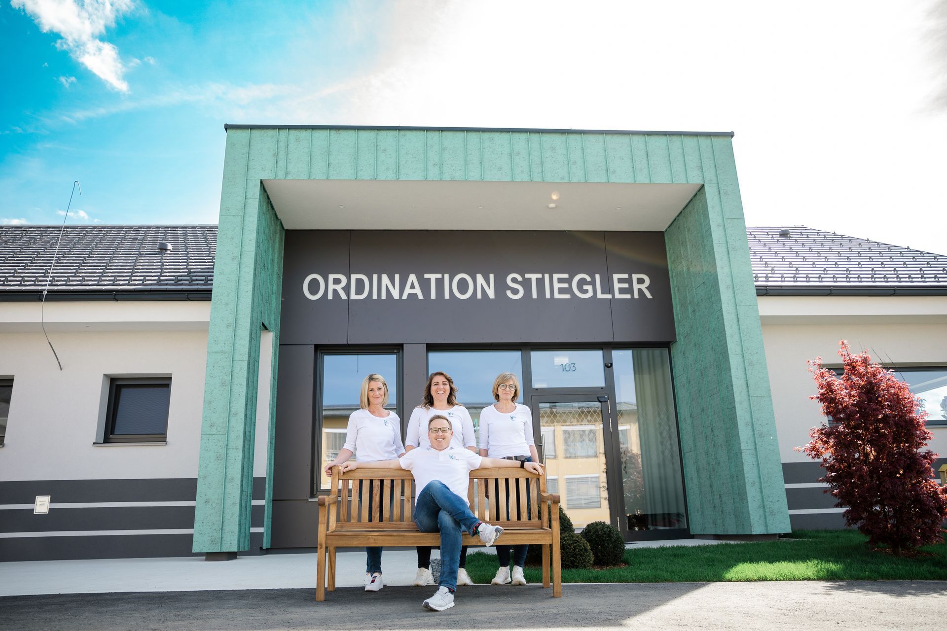 Das Team sitzt auf einer Bank vor einem Gebäude mit der Aufschrift „Ordination Stiegler“