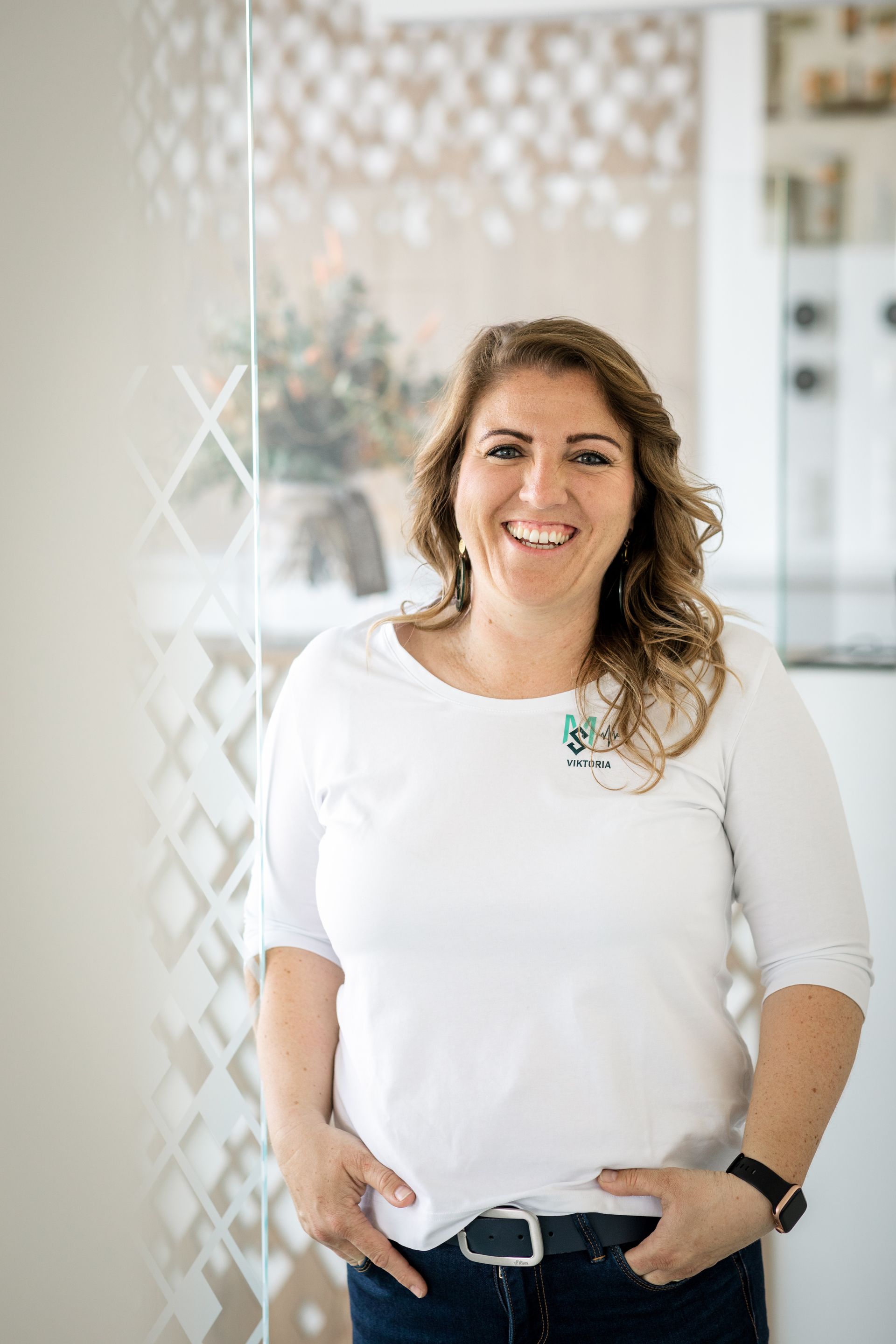 Eine Frau in einem weißen Hemd und blauen Jeans steht mit den Händen in den Taschen da und lächelt.