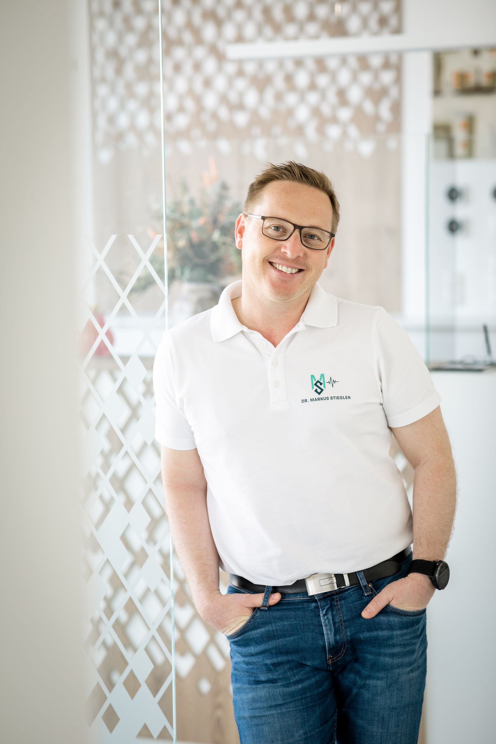 Ein Mann mit Brille und weißem Hemd steht mit den Händen in den Taschen da.