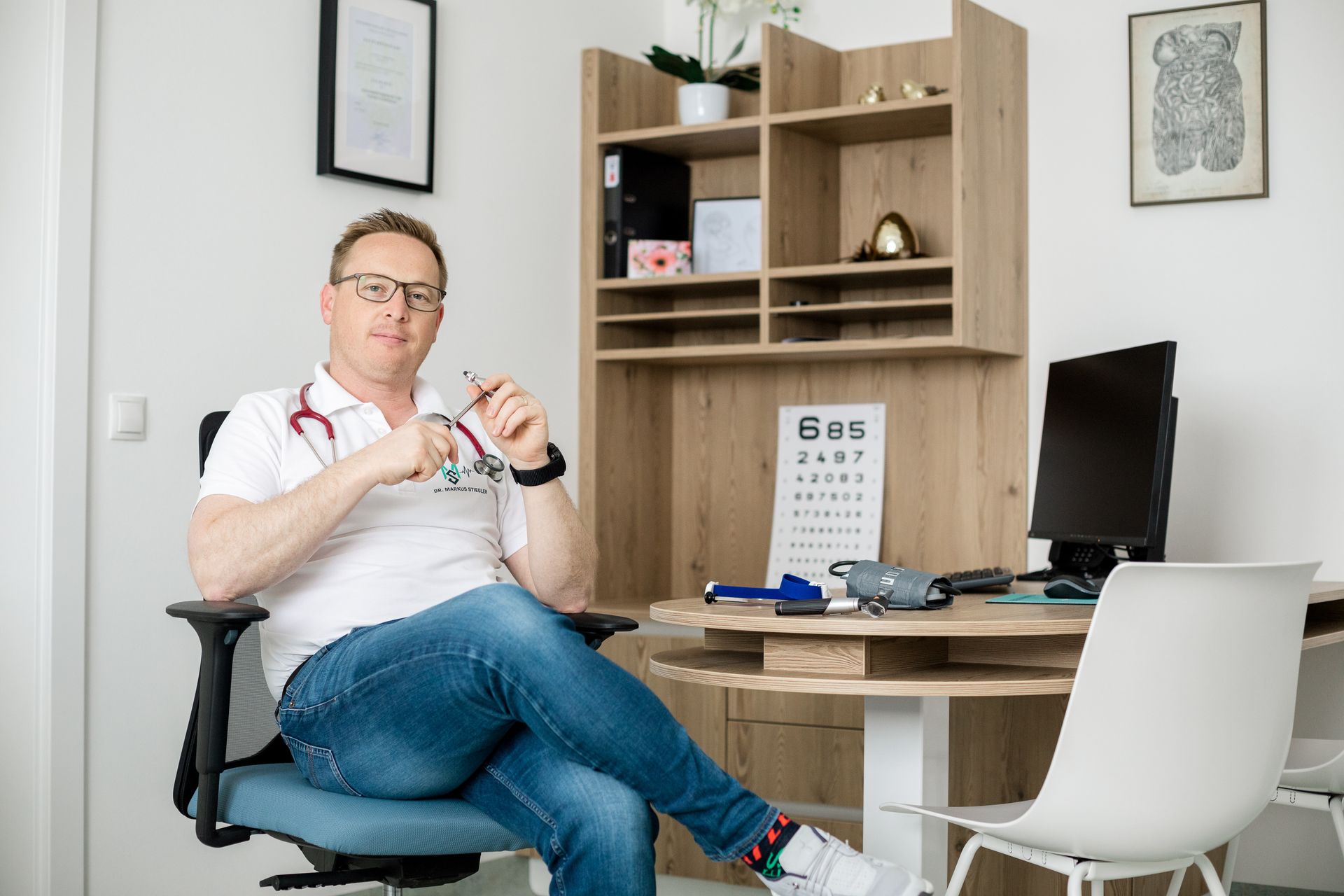 Ein Mann sitzt mit einem Stethoskop um den Hals auf einem Stuhl vor einem Schreibtisch.