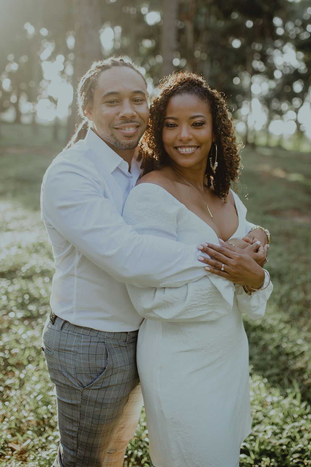 A man and a woman are posing for a picture in a park . the woman is wearing a white dress.