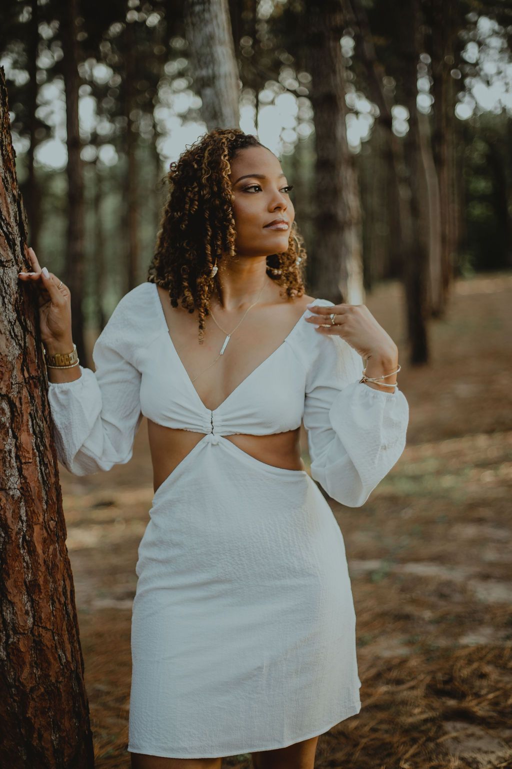 A woman in a white dress is leaning against a tree in the woods.