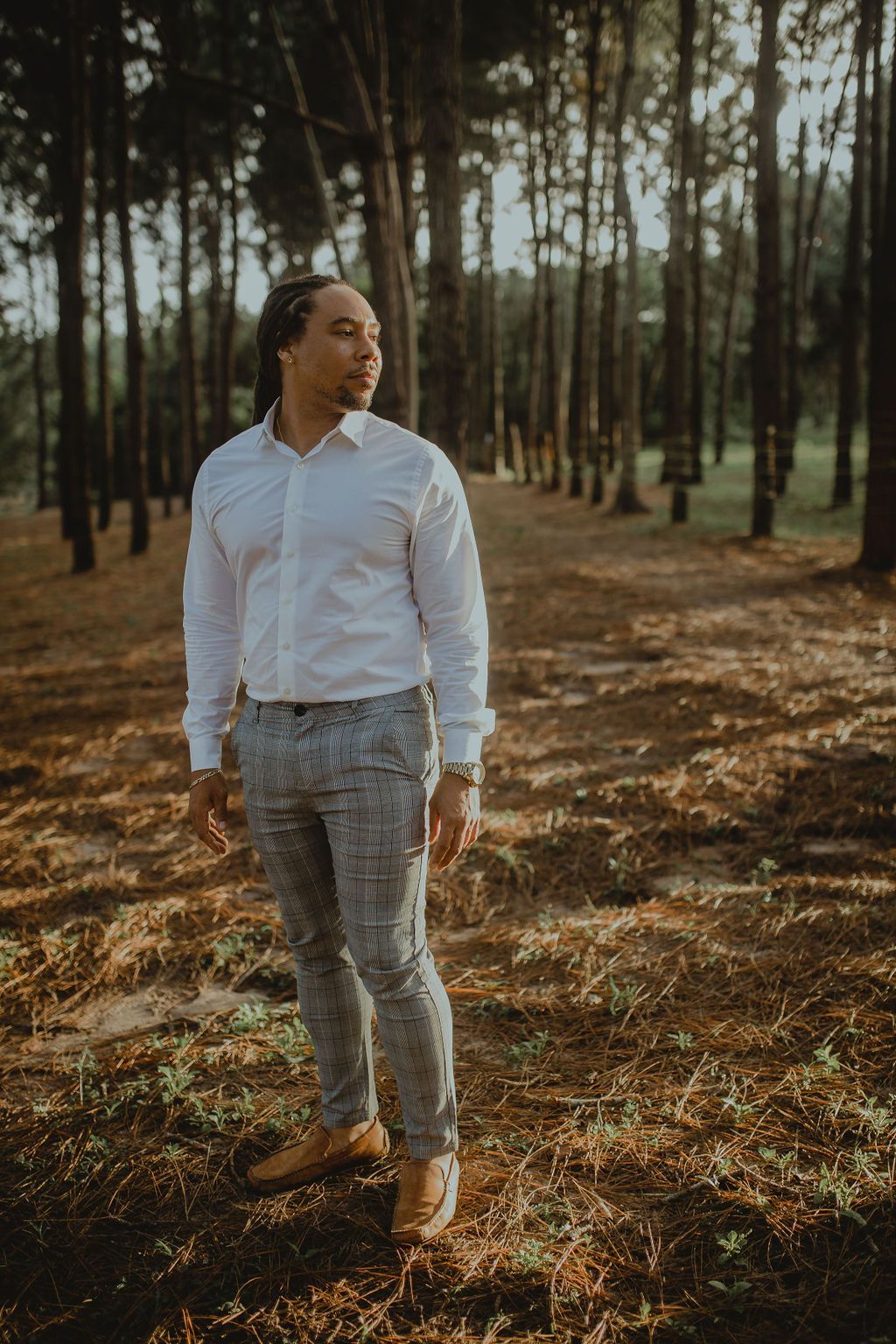 A man in a white shirt and plaid pants is standing in the middle of a forest.