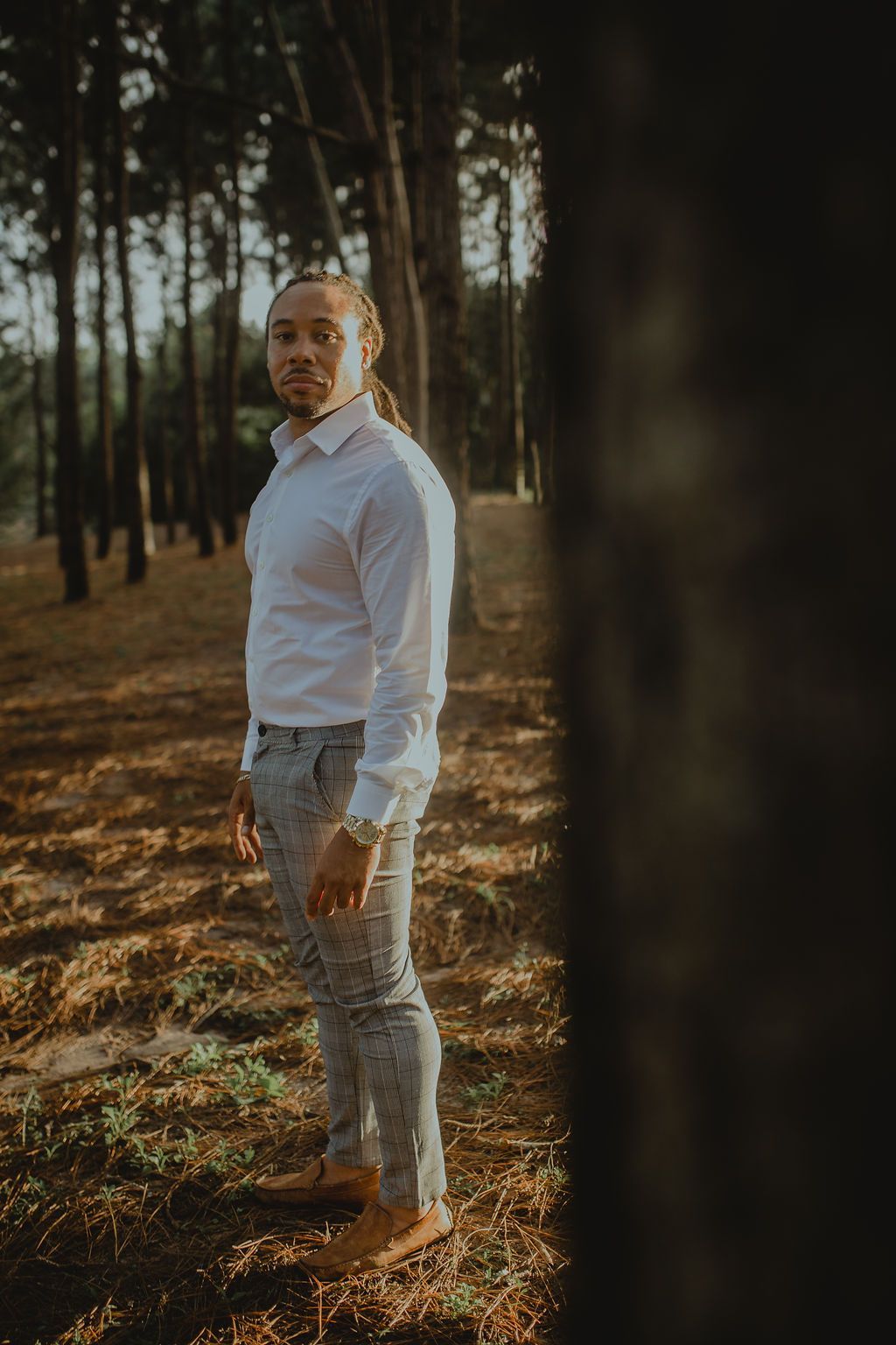 A man in a white shirt and plaid pants is standing in a forest.