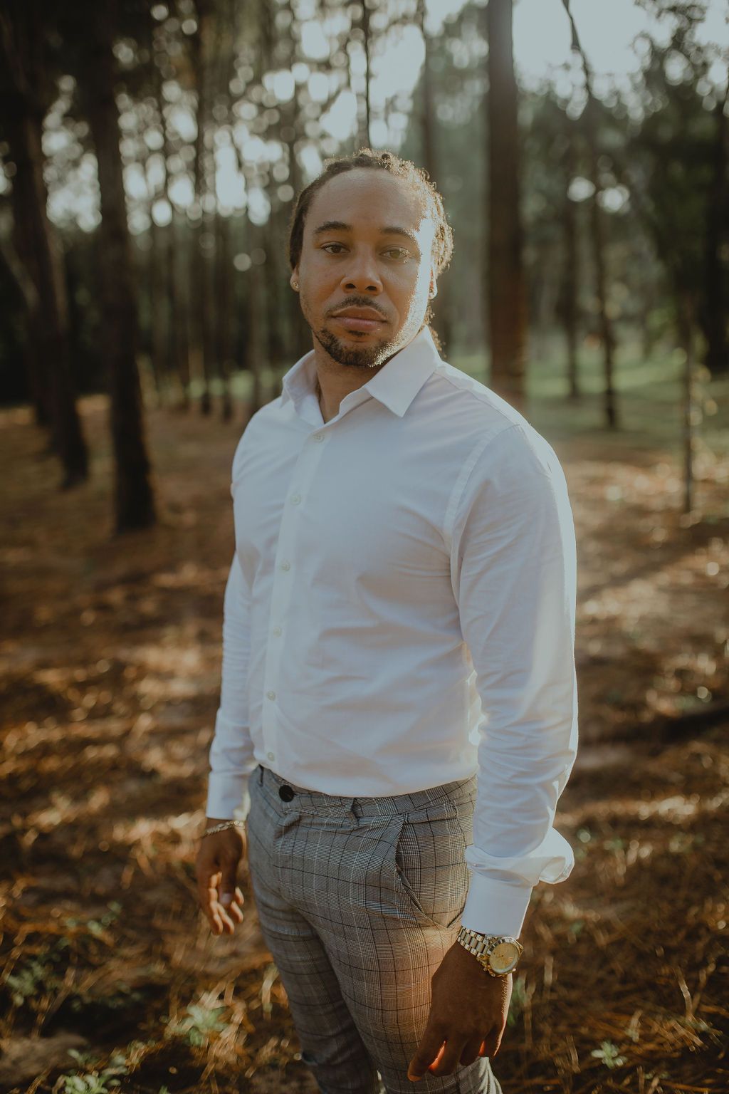 A man in a white shirt and plaid pants is standing in the woods.