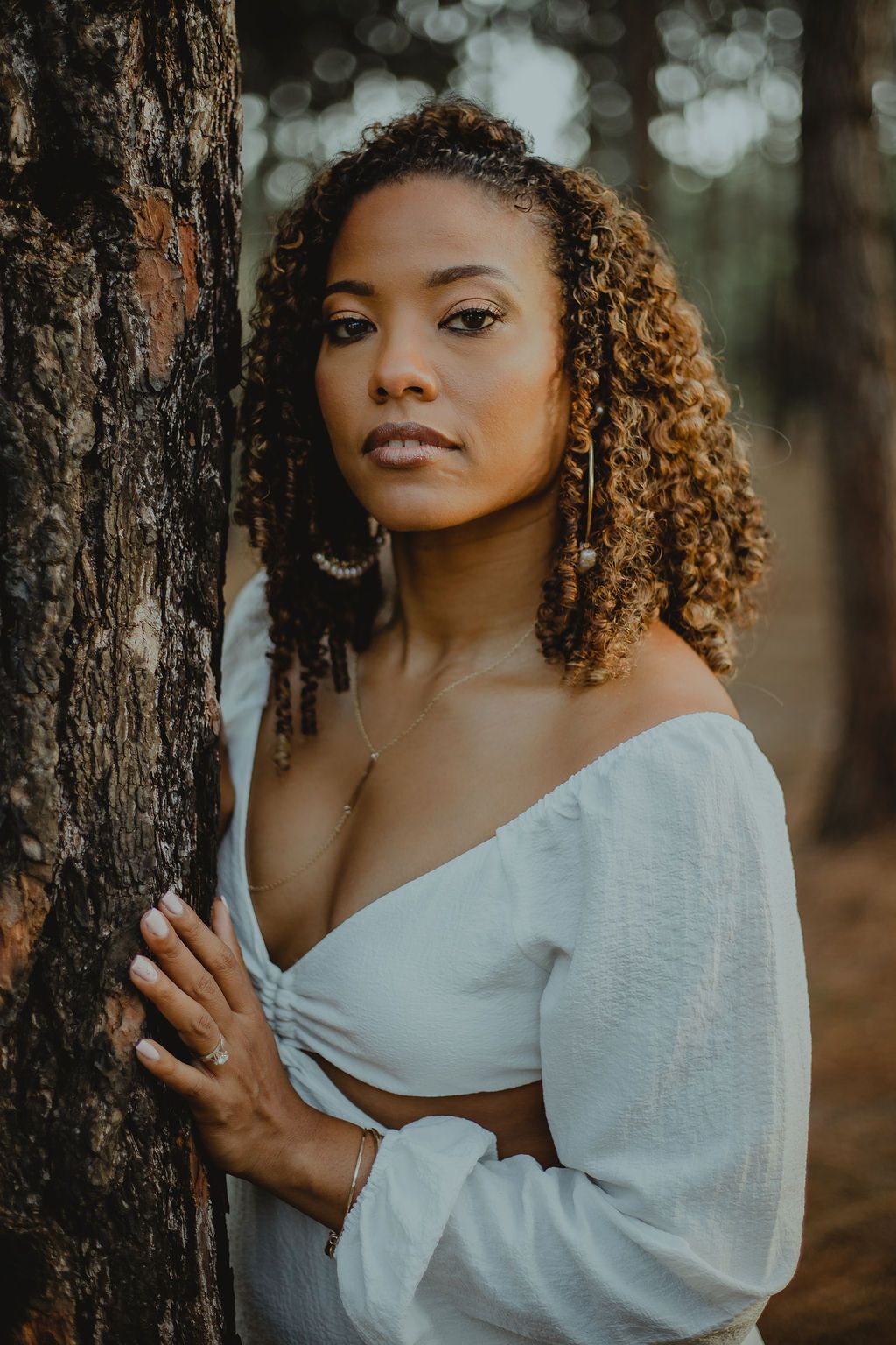 A woman in a white dress is leaning against a tree in the woods.