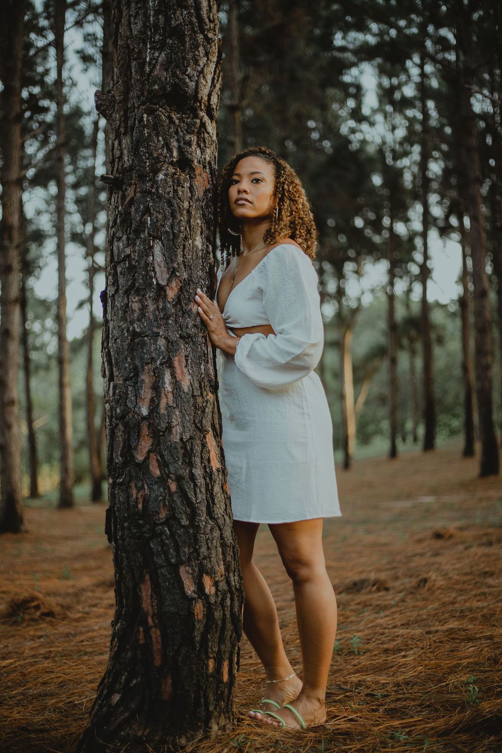 A woman in a white dress is standing next to a tree in the woods.