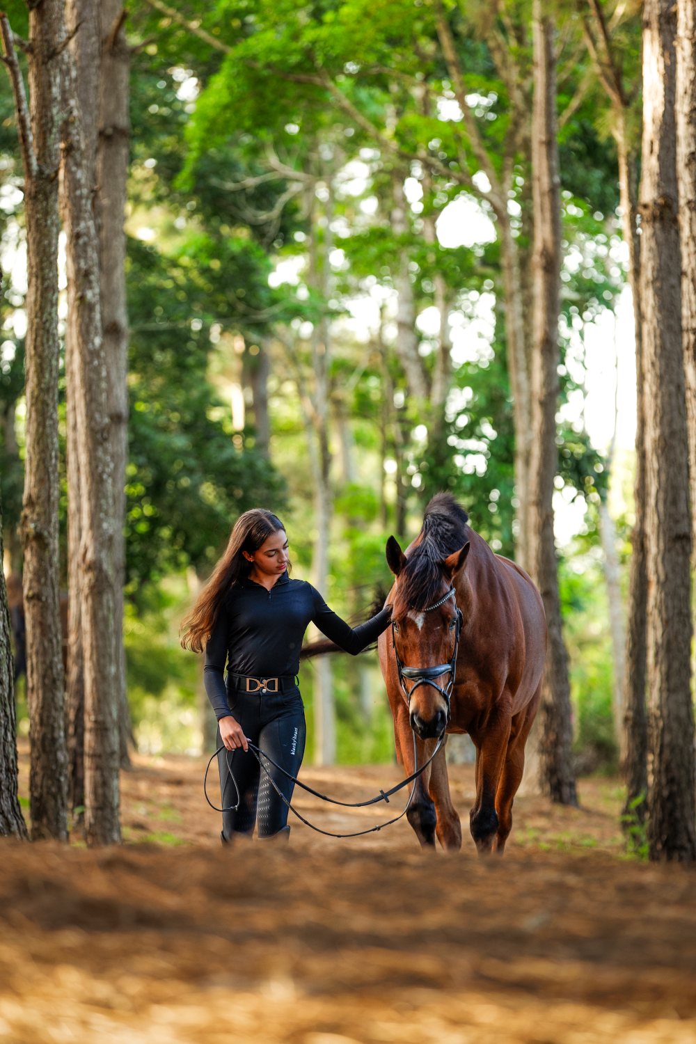 A woman is walking a brown horse through a forest.