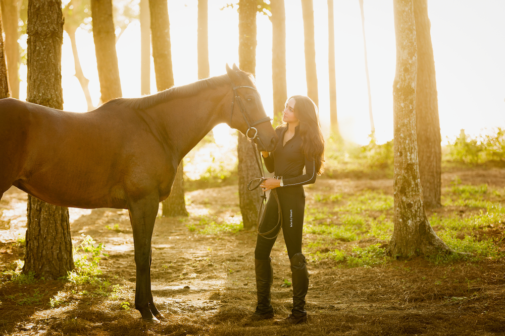 A woman is standing next to a brown horse in the woods.