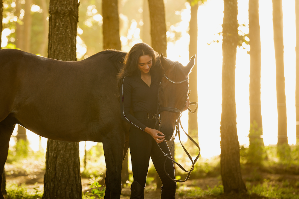 A woman is standing next to a horse in the woods.
