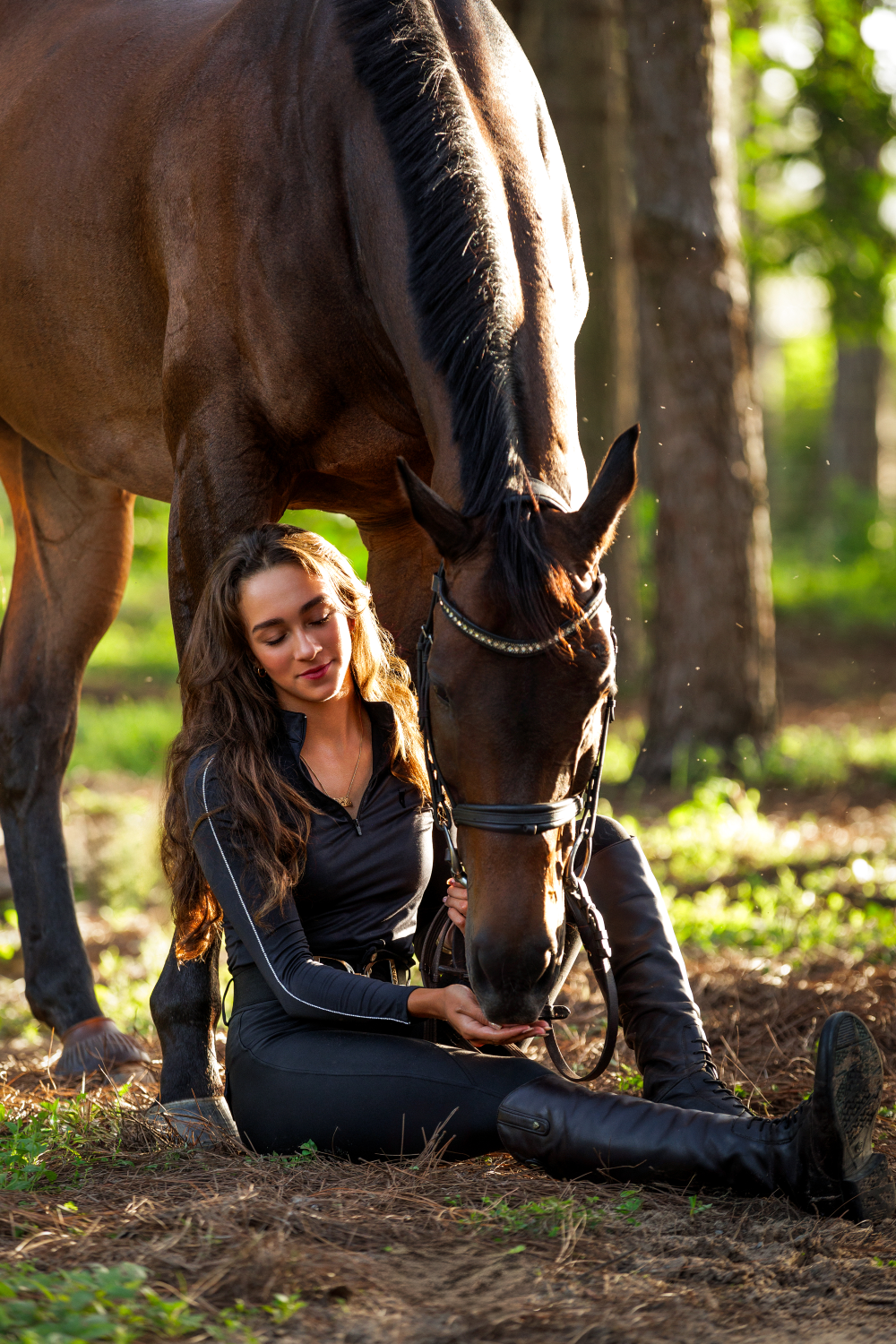 A woman is sitting next to a brown horse in the woods.