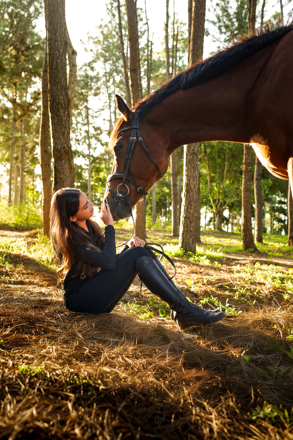 A woman is sitting on the ground next to a horse in the woods.