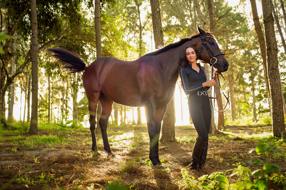 A woman is standing next to a brown horse in the woods.